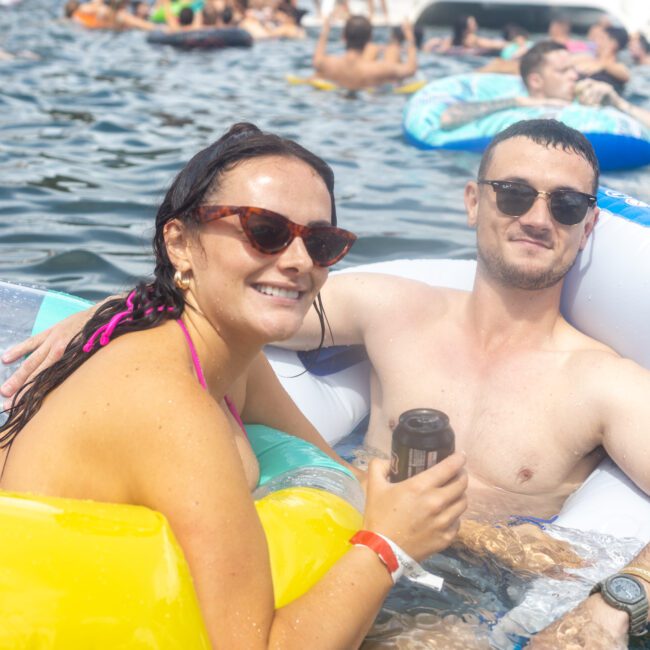 A woman and a man smile while floating on inflatable tubes in a body of water. The woman wears sunglasses and a pink bikini, while the man, also wearing sunglasses, holds a can beverage. Other people are smiling and relaxing in the water on a sunny day, enjoying the fun atmosphere together.