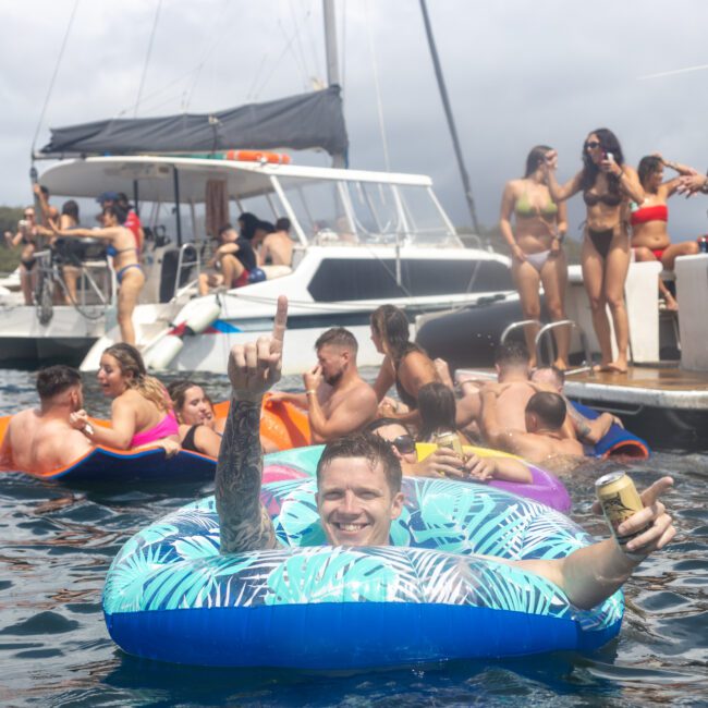 A festive scene at a boat party. People are lounging on inflatables in the water, enjoying drinks, while others socialize on a docked yacht. A man in the foreground smiles and raises a hand in celebration. The atmosphere is lively and relaxed as everyone enjoys the beautiful day on the ocean.