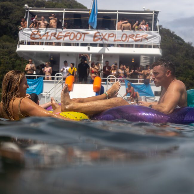 Two people on inflatable rafts float in the water near a large boat named "Barefoot Explorer," with multiple people on the upper deck enjoying themselves. The background boasts lush green trees. A logo reads "YachtSocial.com" in the lower right corner.