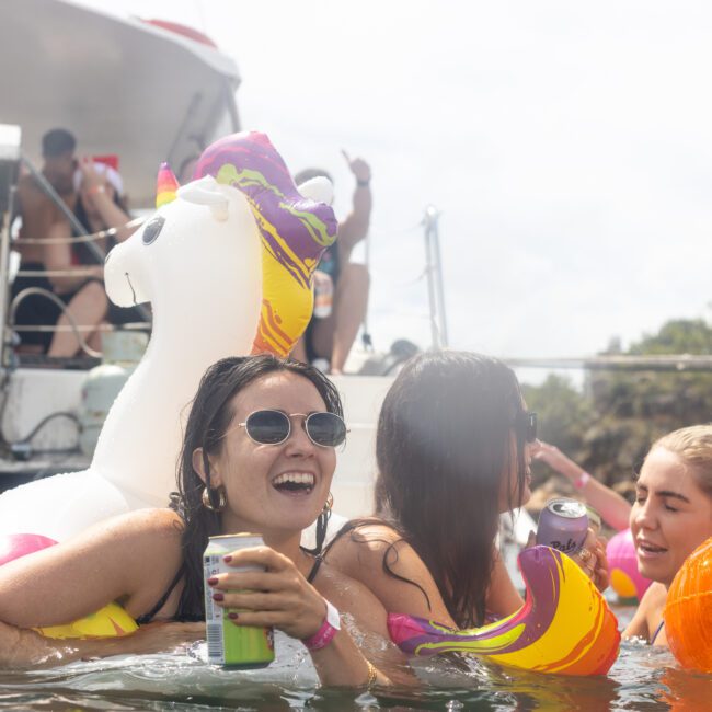 A group of women enjoy a lively boat party, smiling and holding drinks while floating in the water on pool inflatables. One inflatable is shaped like a llama. Other partygoers can be seen on the yacht in the background. The playful logo "Yacht Social Club" is visible in the bottom right.