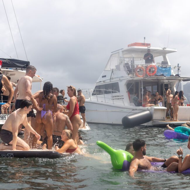 Groups of people relax and have fun on a sunny day on boats and inflatables in the water. Several individuals stand and sit on an anchored float, some in vibrant swimsuits. Two boats are docked nearby, and the city skyline is faintly visible in the background.