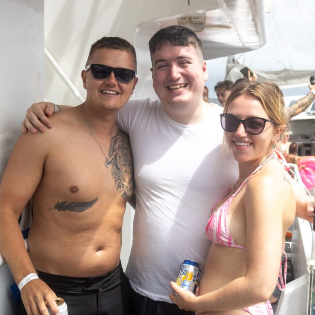 Three people smiling at the camera while standing on a boat during a sunny day. The person on the far left is shirtless, the middle person is wearing a white shirt, and the person on the right is in a pink bikini. They are enjoying their time with others around them.