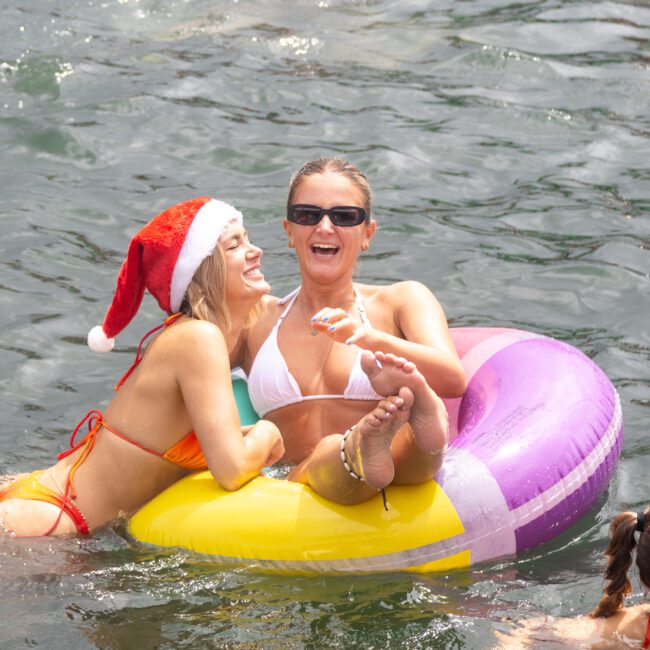 Two smiling women are in a body of water. One, in a red bikini and Santa hat, stands while the other, in a white bikini and sunglasses, floats on a colorful inflatable ring. They laugh under the sun, enjoying their day with others partially visible around them.