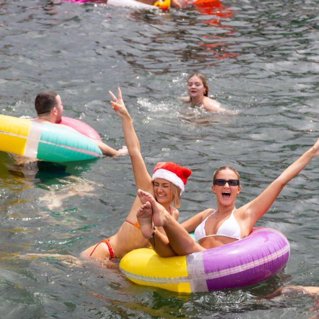 A group of people enjoys a festive time in a body of water, floating on colorful inflatable rings. Two women in the foreground, one wearing a Santa hat, fling their arms in the air, smiling and cheering. Other individuals swim and float around them as laughter fills the air.