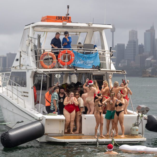 A festive group in swimsuits and Santa hats gathers on a white yacht, docked in a harbor with towering skyscrapers against a cloudy sky. The yacht's upper and lower decks are filled with joyful people celebrating under the city's skyline.