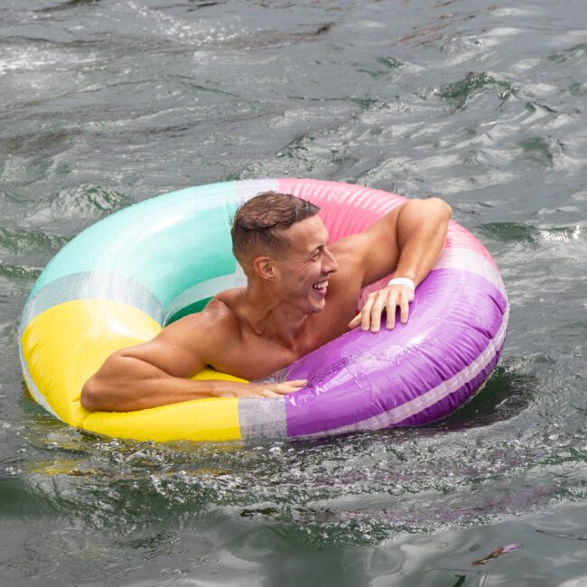 A person with short hair is joyfully floating on a vibrant inflatable ring in a body of water. They are smiling widely and gripping the sides of the ring. The water is slightly wavy around them, enhancing the fun atmosphere.