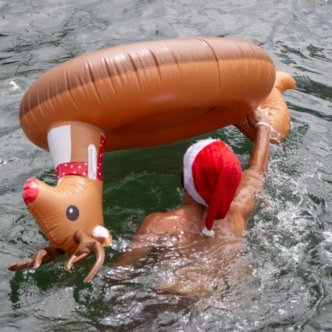 A person wearing a Santa hat swims in a body of water with a reindeer-shaped inflatable ring, complete with a red nose and antlers, enhancing the festive holiday theme. The water is greenish and appears to be moving gently under the clear sky.