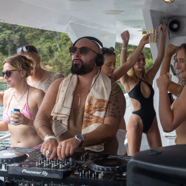 A group of people are partying on a boat. A man wearing sunglasses, headphones, and a towel around his neck is DJing at a turntable, setting the perfect vibe. Several women in swimwear dance and hold drinks. Lush greenery and sparkling water are visible in the background, creating an idyllic summer scene.