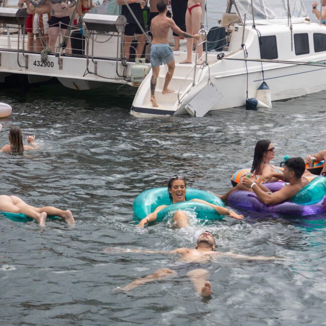 A group of friends enjoy a sunny day on the water, floating on vibrant inflatable rings and swimming near a docked boat. Some lounge on the inflatables while others swim around, laughing and having fun. The boat's registration number is visible, making it easy to identify.