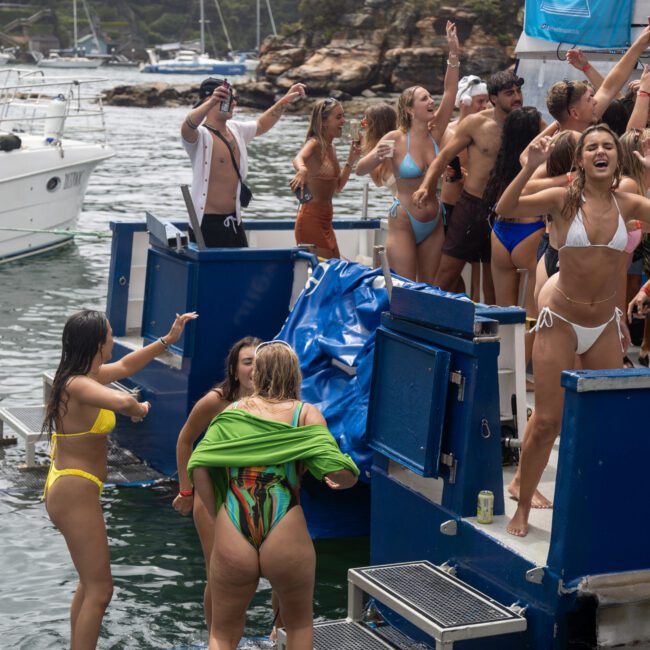 A group of people is enjoying a lively party on a boat. Some are dancing and cheering, while others are mingling. Most are in swimwear, and a couple of people are swimming nearby. Another boat is visible in the background, docked against a picturesque shoreline under the clear sky.