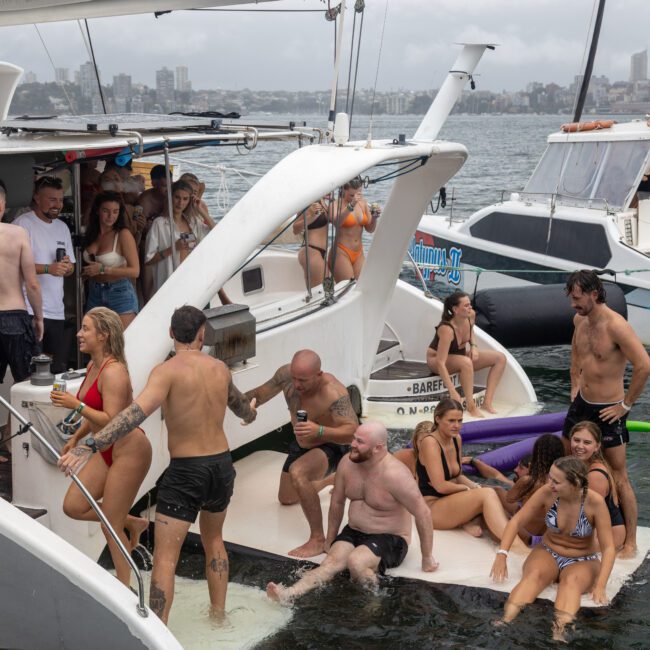 A lively scene on a large sailboat shows people enjoying a party. Men and women in swimwear are socializing, swimming, and dancing. A nearby sailboat adds to the festive atmosphere. The background features a picturesque waterfront cityscape under a cloudy sky.