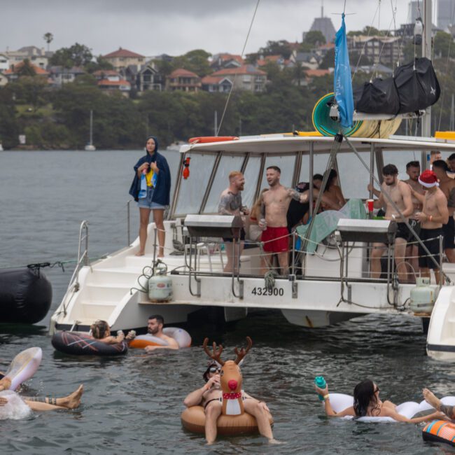 A group of friends enjoying a lively celebration on a catamaran anchored in a picturesque bay. Some are on the boat, while others float nearby on inflatables, including a reindeer. The sky is overcast, and charming shoreline houses and trees create a beautiful backdrop.