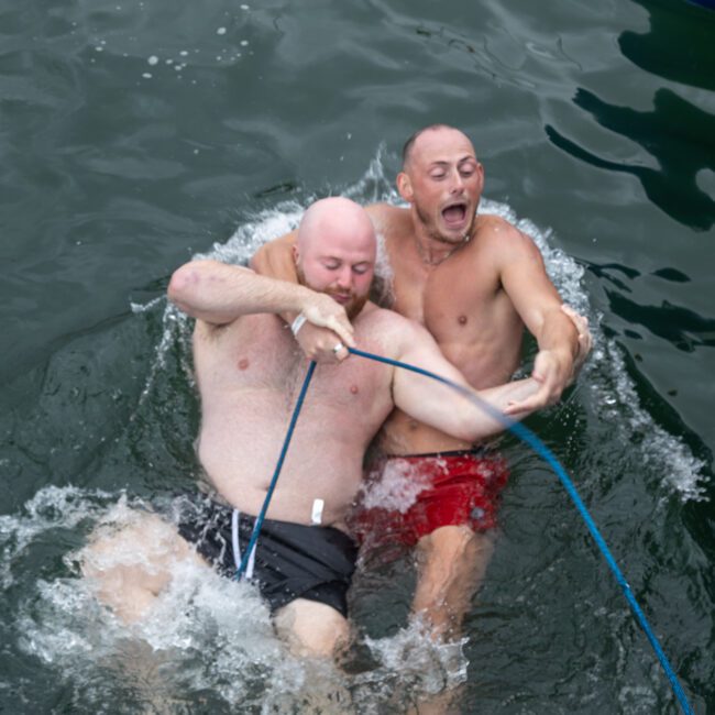 Two shirtless men are seen wrestling and holding onto a blue rope while splashing in the water. The man on the left is bald with a beard, and the man on the right has short hair and appears to be yelling, adding excitement to their playful moment. Both are wearing swim trunks and seem to be enjoying themselves.