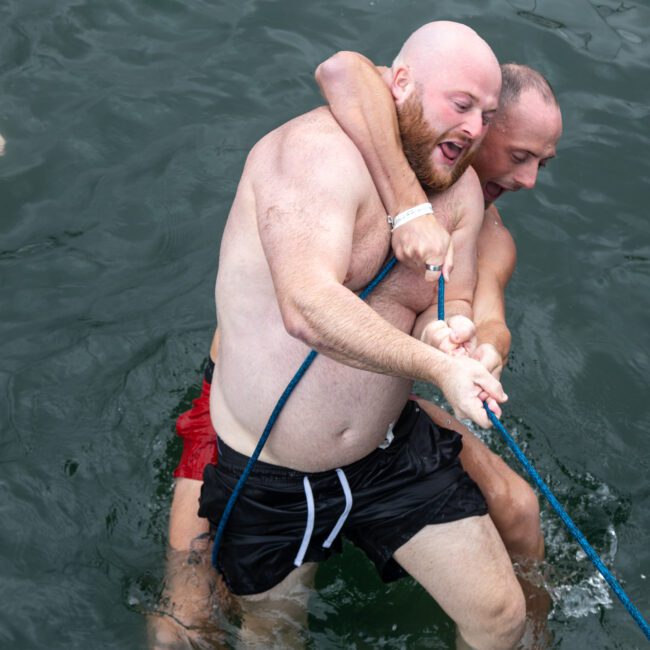 Two shirtless men are playfully struggling in the water while holding onto a blue rope, engaging in a friendly game. One man has his arms around the other, both smiling and laughing, creating a lighthearted and fun atmosphere.