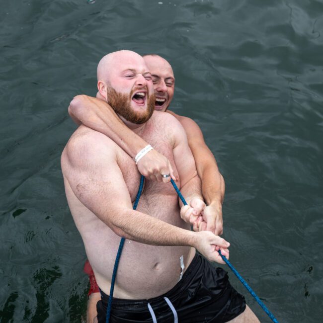 Two bald men, one with a beard and the other bare-faced, enjoy a playful moment in water. Both shirtless, they struggle over a vibrant blue rope. Laughing in pure joy, the scene perfectly captures their competitive spirit against the backdrop of rippling water’s surface.