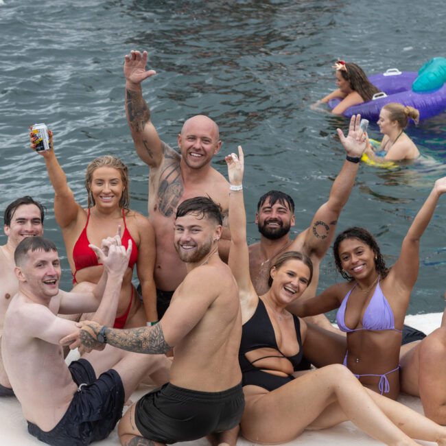 A group of ten people are sitting and kneeling on a floating platform in the water, smiling and raising their arms. They are in swimsuits, enjoying a sunny day. In the background, a person floats on an inflatable unicorn while others swim cheerfully. The scene exudes pure joy and togetherness.