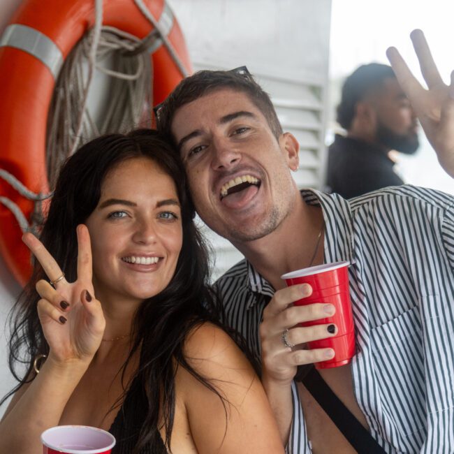 A woman and a man smile at the camera while holding red cups and making peace signs with their hands. They are standing on a boat deck with an orange life preserver visible in the background. The atmosphere appears relaxed and festive, highlighting a joyful moment on the water.