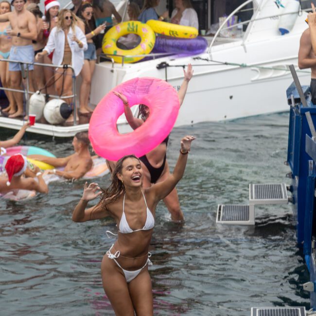 A group of friends enjoy a boat party, with some in the water holding inflatable rings. A woman in the foreground wearing a white bikini raises her arm while holding a pink flamingo ring, smiling. Boats and more people are seen in the background having fun.