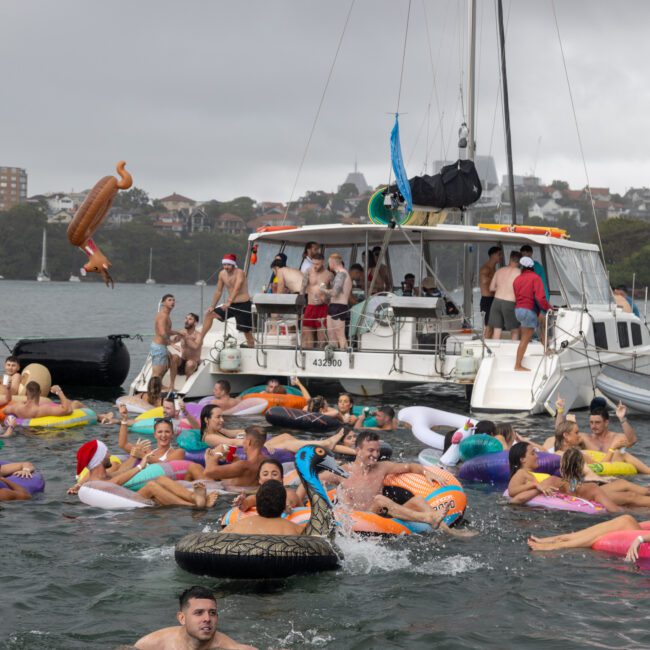 People are swimming and floating on various inflatable toys near anchored boats at a lively festive gathering on the water. The scene is cheerful, with individuals wearing swimsuits and holiday-themed accessories. The background shows a partly cloudy sky and charming coastal buildings.