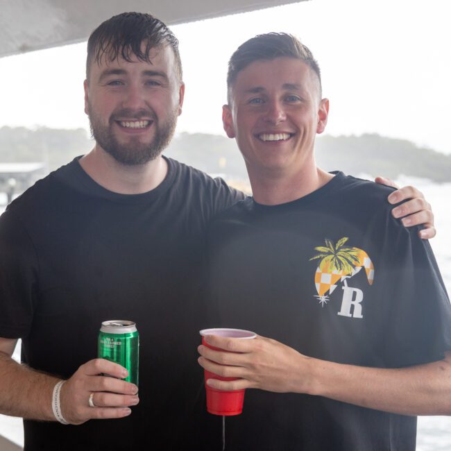 Two smiling men stand close together on a boat, holding drinks—a green can and a red cup. One has his arm around the other. Dressed in casual dark shirts, they exude camaraderie against a backdrop of water and land, creating an atmosphere of joy and relaxation.