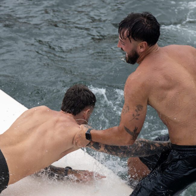 Two shirtless men are playfully wrestling on the edge of a floating platform or boat, with one man grabbing the other's arm. The scene is set on water, and both appear wet, suggesting they have been swimming. The background shows sparkling ripples.