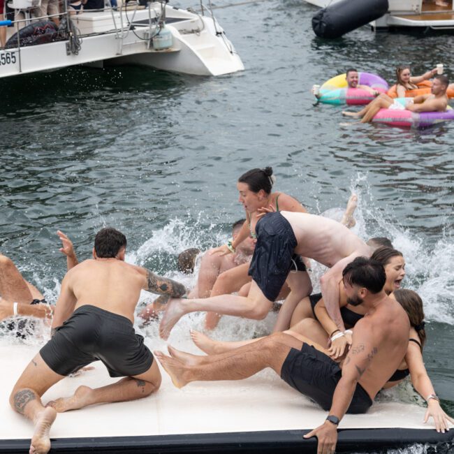 A group of people in swimsuits is playfully falling into the water from a floating platform next to a docked boat. Some individuals are seen on inflatable tubes in the background. The scene is lively and spirited, capturing a fun water activity, typical of events like Boat Parties Sydney by The Yacht Social Club.