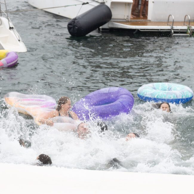 A group of people in a lake splashing water and having fun on colorful inflatable tubes, enjoying the carefree summer day. Some boats are docked in the background.