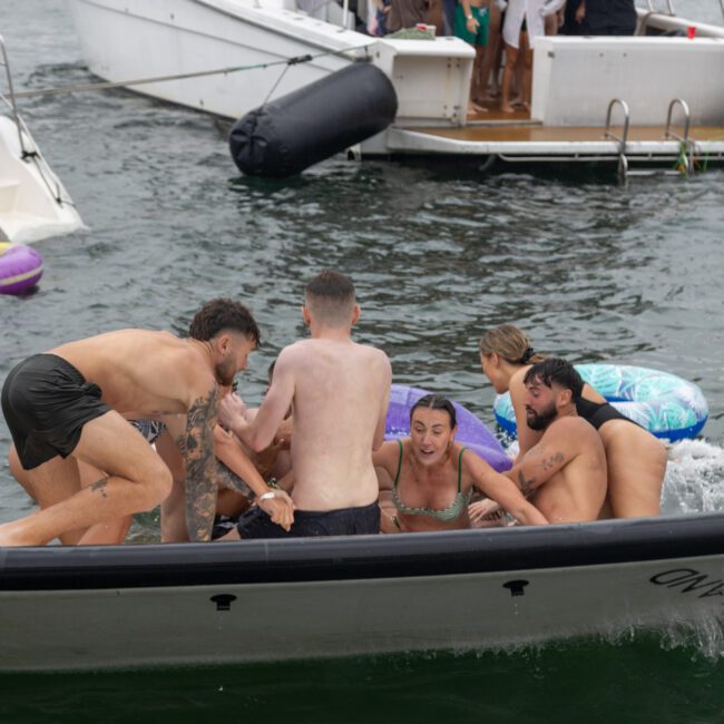 A lively group of young people are playing on and around an inflatable boat in the water. Some are in swimwear, laughing and holding onto the boat. Other boats float in the background with more people relaxing and watching under the warm sun.