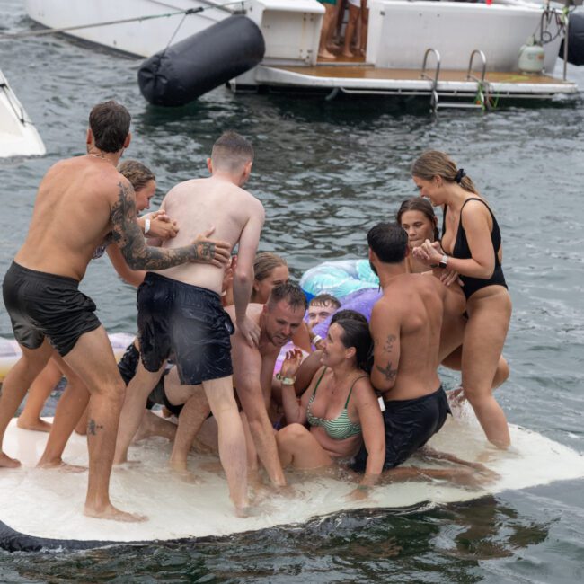 A group of people, wearing swimsuits, try to balance on a floating platform in the water. Some are standing while others are sitting or kneeling. Boats and colorful inflatables in the background suggest they're enjoying an adventurous day at sea or a lake.