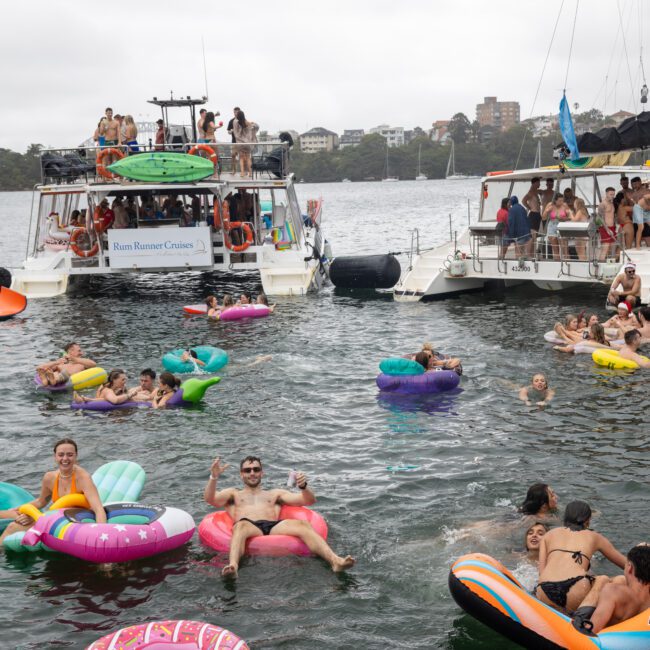 A lively scene featuring people enjoying themselves in a body of water, surrounded by floating boats and inflatables. Various individuals, including children and adults, are swimming, lounging on floaties like flamingos and unicorns, and socializing around several anchored boats.