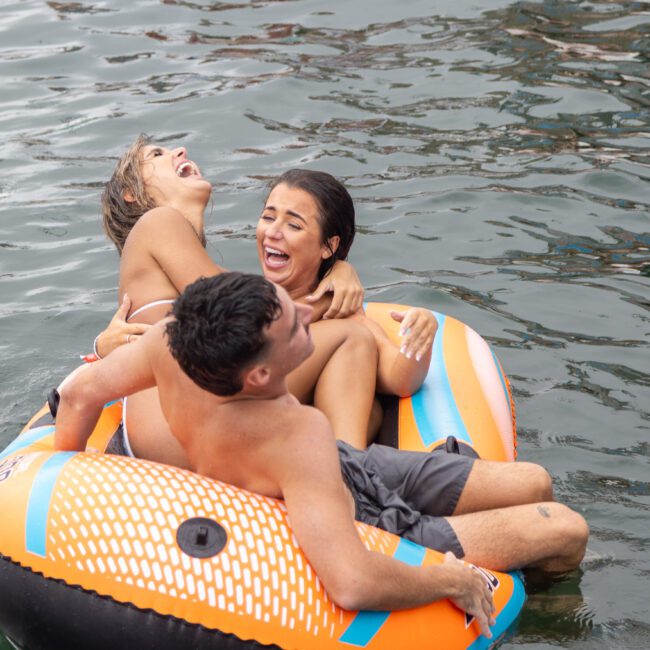 Three people are laughing and having fun on an inflatable raft in the water. One person is leaning back, while another holds onto them, and the third person sits in the raft, all sharing a lively moment of adventure together.