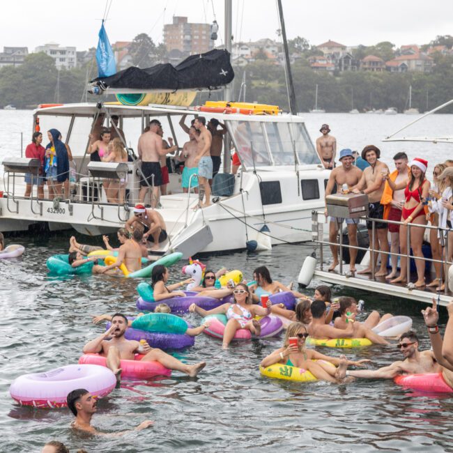 A lively scene of people enjoying a party on the water, surrounded by boats and colorful inflatables. The weather seems overcast but the atmosphere is cheerful. Many are wearing swimsuits, and some boats display small blue flags fluttering in the breeze as everyone celebrates together.