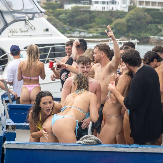 A group of people in swimwear are partying on a boat and a dock next to another boat. Some are dancing, drinking, and talking. The scene is lively with The Yacht Social Club Sydney Boat Hire, as everyone enjoys themselves near the water.