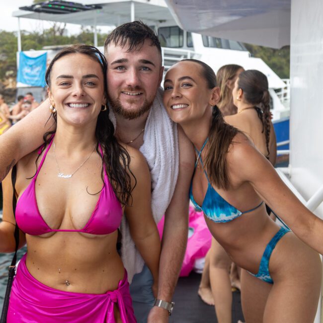 Three people smile and pose for a photo on a boat during a lively party. Two women in bright bikinis stand on either side of a man in a white towel. In the background, you can see an inflatable slide and other party-goers enjoying the sunny day.