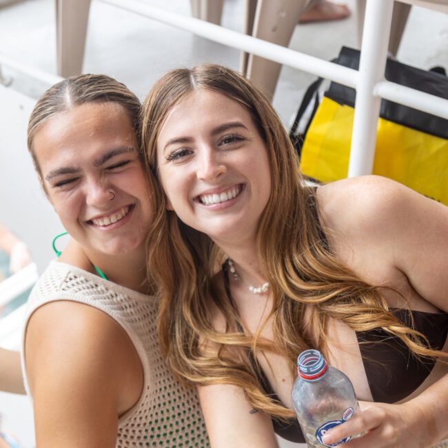 Two women are smiling at the camera while onboard a boat, enjoying time together. One woman in a vibrant crochet top is holding a water bottle, and the other in a brown top is also smiling widely. People and part of the boat's modern structure are visible in the background.