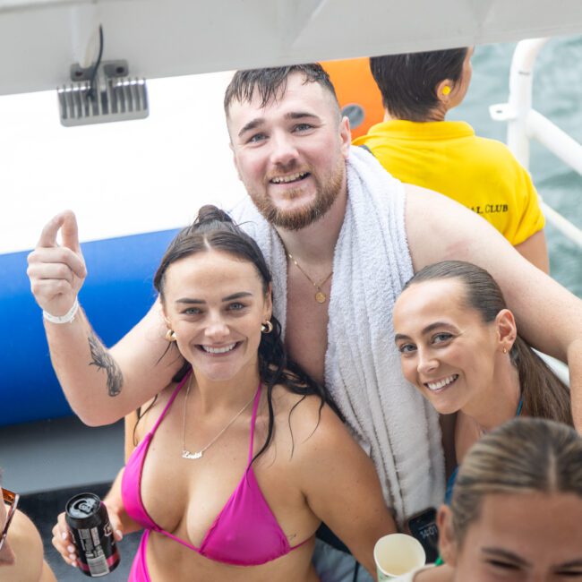 Three people are smiling and posing together on a boat. The person in the center has a towel draped over his shoulders, and the two people on either side are wearing swimwear. Other passengers and the beautiful ocean are partially visible around them.