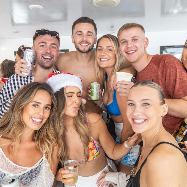 A group of eight young adults is posing happily for a photo on a boat. They are dressed in swimsuits and casual summer clothing, with one person wearing a Santa hat. Some are holding drinks and smiling brightly, creating a festive and lively atmosphere under the sun.