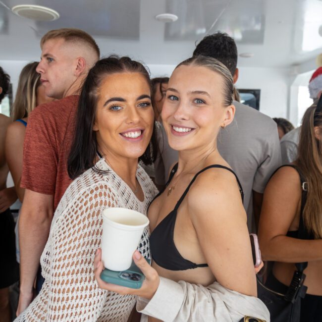 A group of people are gathered at a lively indoor event. Two women in the foreground are smiling at the camera, one holding a cup. The atmosphere is cheerful, with others in the background engaging in conversations and activities. Some attendees are adding a touch of fun by wearing beachwear.