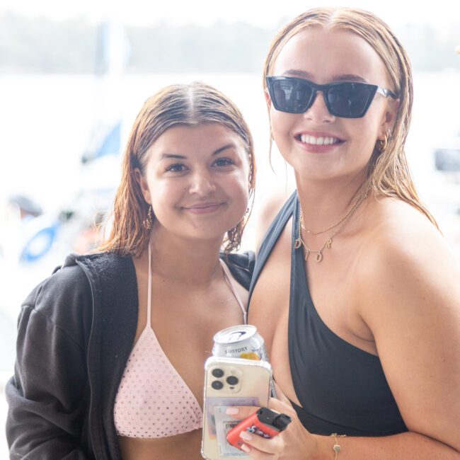 Two women stand close together, smiling at the camera. One wears a black hoodie over a light pink bikini, holding a phone and a drink, while the other sports sunglasses and a black swimsuit, also with a phone. They are outdoors near sparkling water, enjoying the sun.