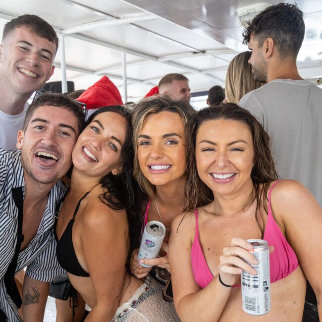 A group of five young adults smiles at the camera while on a boat. Four women, dressed in swimsuits, hold canned drinks. A man in a striped shirt joins them, smiling. Other people and boat structures are visible in the background, showcasing the lively atmosphere of The Yacht Social Club Sydney Boat Hire.