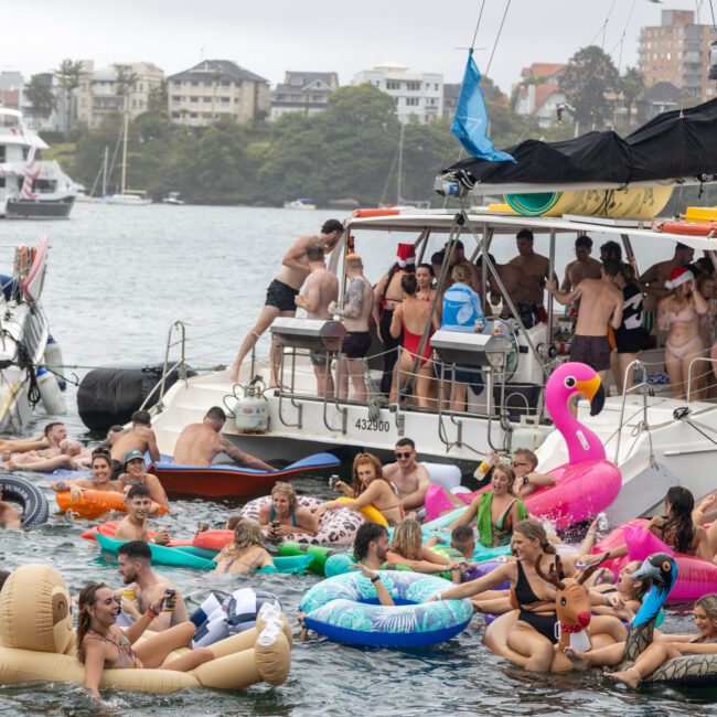 A lively scene of people partying on a boat and floating in a body of water. Numerous individuals are on various inflatables such as flamingos, doughnuts, and pool noodles. In the background, you can see palm trees swaying gently in the breeze and additional boats. Everyone appears to be having fun.