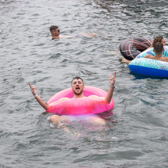 A man in a pink inflatable ring is in the water, playfully showing a peace sign with both hands. Around him, three other people are lounging on various brightly colored inflatable tubes, enjoying a delightful water activity. The day appears cloudy and the water is calm.