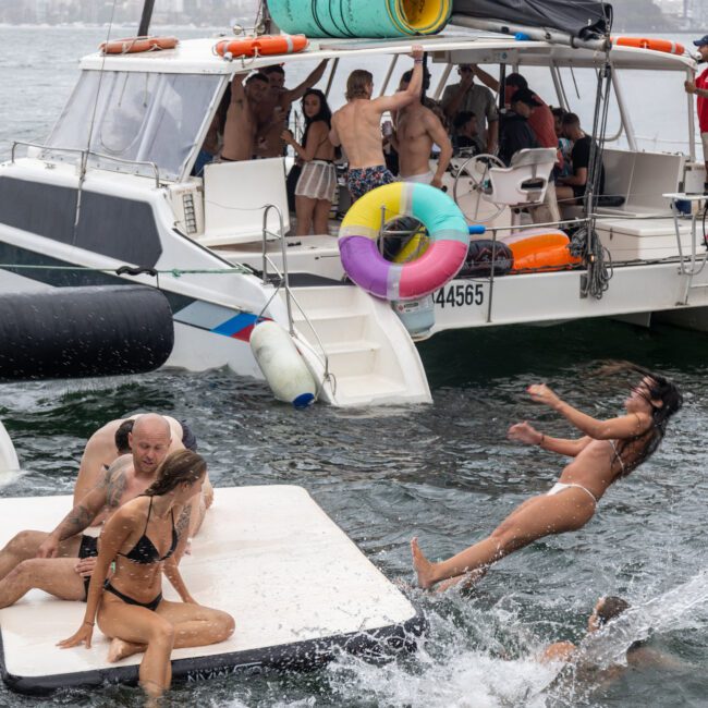 Group of people enjoying a day on the water with some on a boat and others on a floating platform. Two women in bikinis sit on the platform, while another woman is mid-air, jumping into the water. The boat is equipped with colorful inflatables and features fun games for everyone.