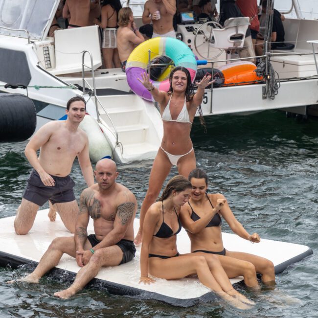 A group of friends in swimwear are having fun on a floating platform next to a boat. Some are sitting and posing for the camera, while a woman in a white bikini stands holding up an inflatable ring. The boat in the background is filled with more people and colorful inflatables.