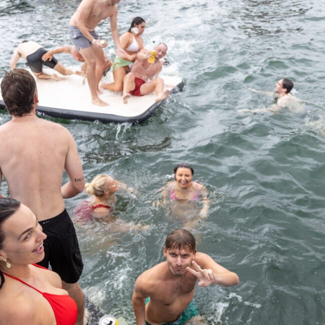 A group of people enjoy a pool party in the water. Several individuals float on inflatable platforms, swim, smile, and wave at the camera. Some drinks are held as people engage in lively, playful activities, with boats visible in the background and laughter filling the air.