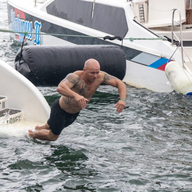 A man with tattoos and a shaved head is diving into the water from a boat named "Barefoot II." He is wearing black shorts. In the background, there are other boats, kayaks, and inflatables in the water, creating a lively and recreational scene.