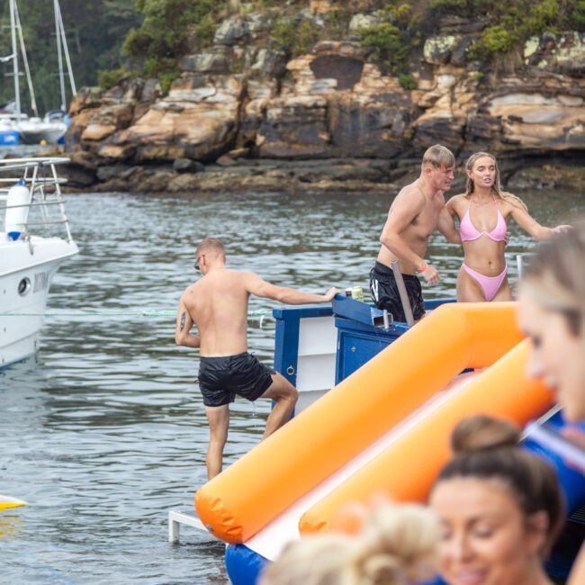 A group of people enjoys a day out on the water near a rocky shore. Some are on a boat, while others use an inflatable slide to enter the water. A man climbs a ladder onto the boat, and a woman in a pink bikini stands nearby, taking in the scenery. Other boats are visible in the background.