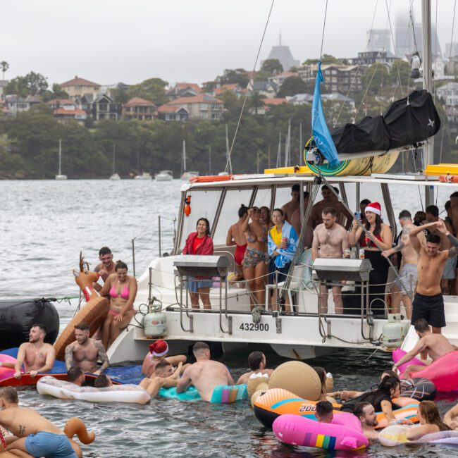 A lively scene of people enjoying a party on and around a boat. Many are in swimsuits, with some dancing on the deck and others floating in the water on colorful inflatables shaped like flamingos, unicorns, and other animals. The city skyline adds to the vibrant atmosphere.