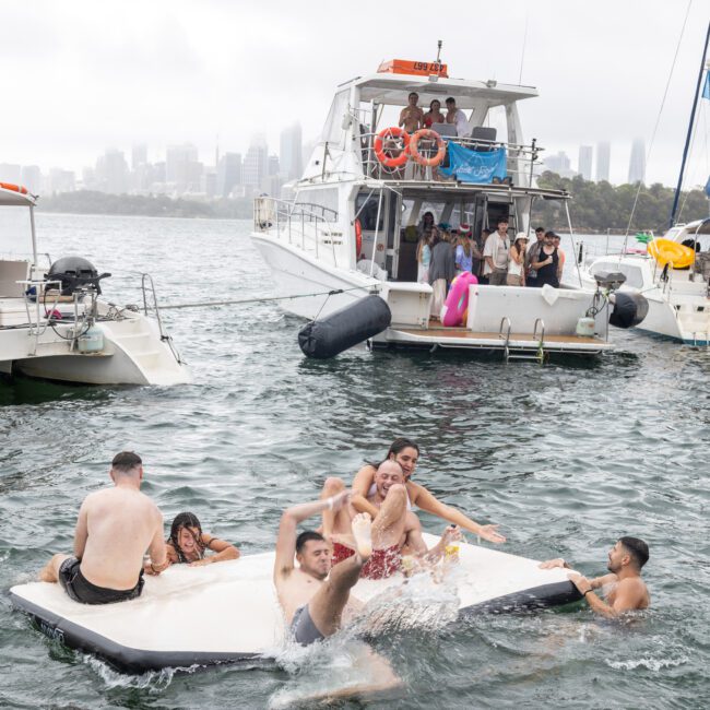 People are enjoying a day on the water with several yachts anchored nearby. Some individuals are playing on a floating raft, while others relax on the yachts. The city skyline is visible in the background under a cloudy sky.