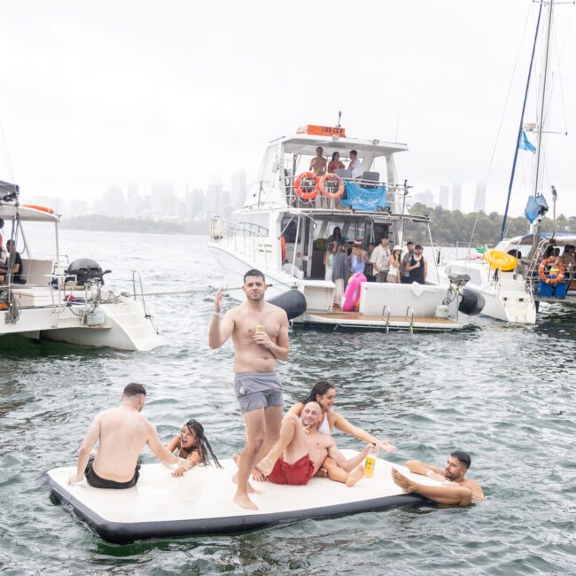 A group of people enjoys a party on anchored boats in a large body of water. Some are on the boats while others relax on a floating mat, surrounded by colorful inflatables. They seem to be having fun with the city skyline faintly visible in the foggy distance, creating an enchanting atmosphere.